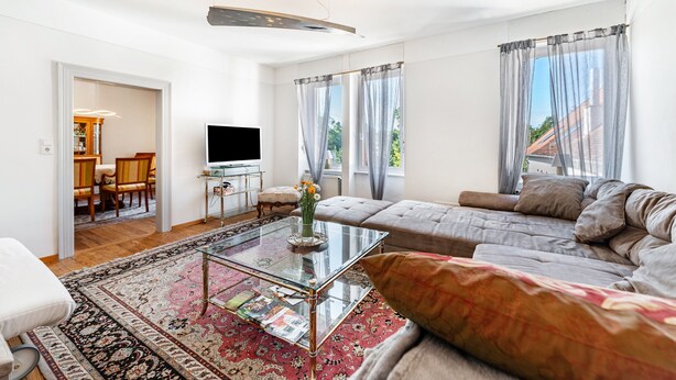 A modern living room with a sofa, a glass coffee table, a flat-screen TV, and a view of the outdoors through large windows.