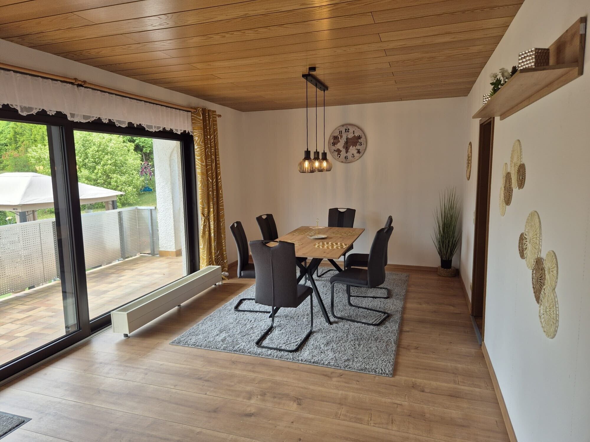 A dining area with a wooden table and chairs, a large window with curtains, a wall clock, and hanging pendant lights.