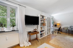 A living room with a television, bookshelf, and a window with plants.