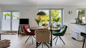 Ett modernt matområde med ett runt bord, stolar, en tv och utsikt över omgivningen genom stora fönster.