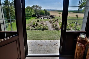 A view from inside a room through a glass door, looking out at a circular garden with a central structure, surrounded by greenery and a clear sky.