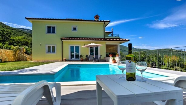 Una casa de dos pisos con piscina, mobiliario exterior y una botella de vino sobre una mesa.