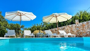 Piscina con sillones blancos y sombrillas, rodeada por un muro de piedra y follaje.