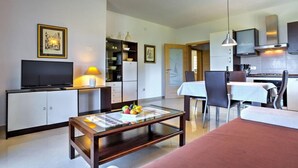 A modern living room with a flat-screen TV, a glass coffee table with fruit, a sofa, a dining area with a table and chairs, and a kitchen with cabinets and a microwave.