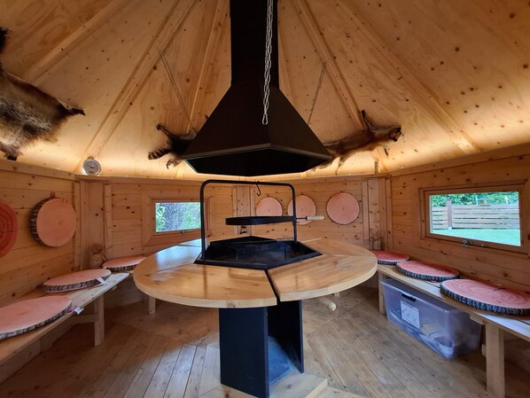A wooden cabin interior with a central table, benches, and a chimney.