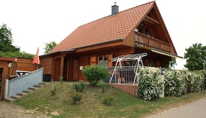 A wooden house with a sloped roof, a balcony, and a small garden with white flowers.