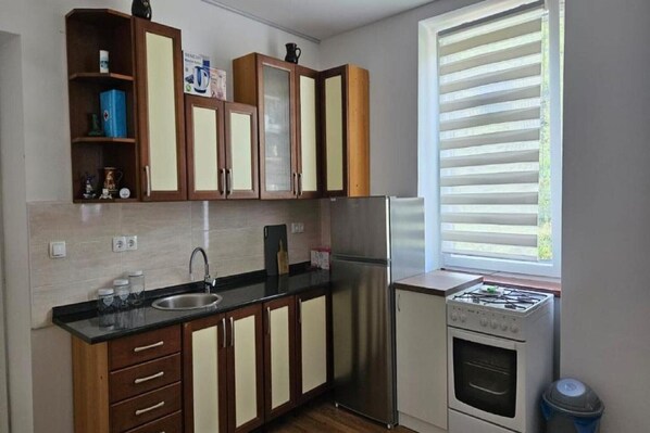 A compact kitchen with wooden cabinets, a stainless steel refrigerator, a gas stove, and a window with blinds.