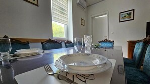 A dining area with a table set for two, featuring a white tablecloth, two plates, a glass, and cutlery. The room has blue upholstered chairs, a window with blinds, and a door. - Apartment Gracac - Two Bedroom Apartment with Terrace (Gracac)