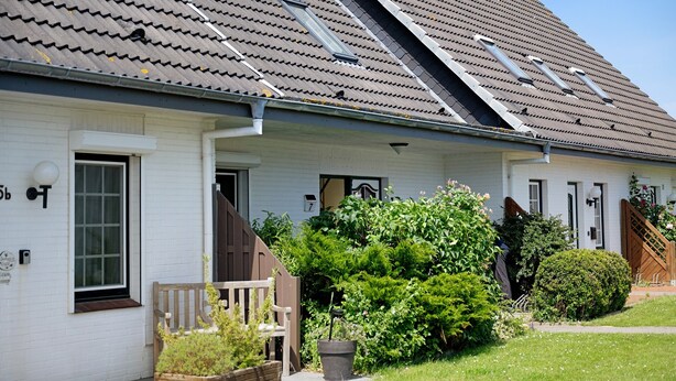 En hvid bygning med skråtag, en træbænk og en potteplante i forhaven.