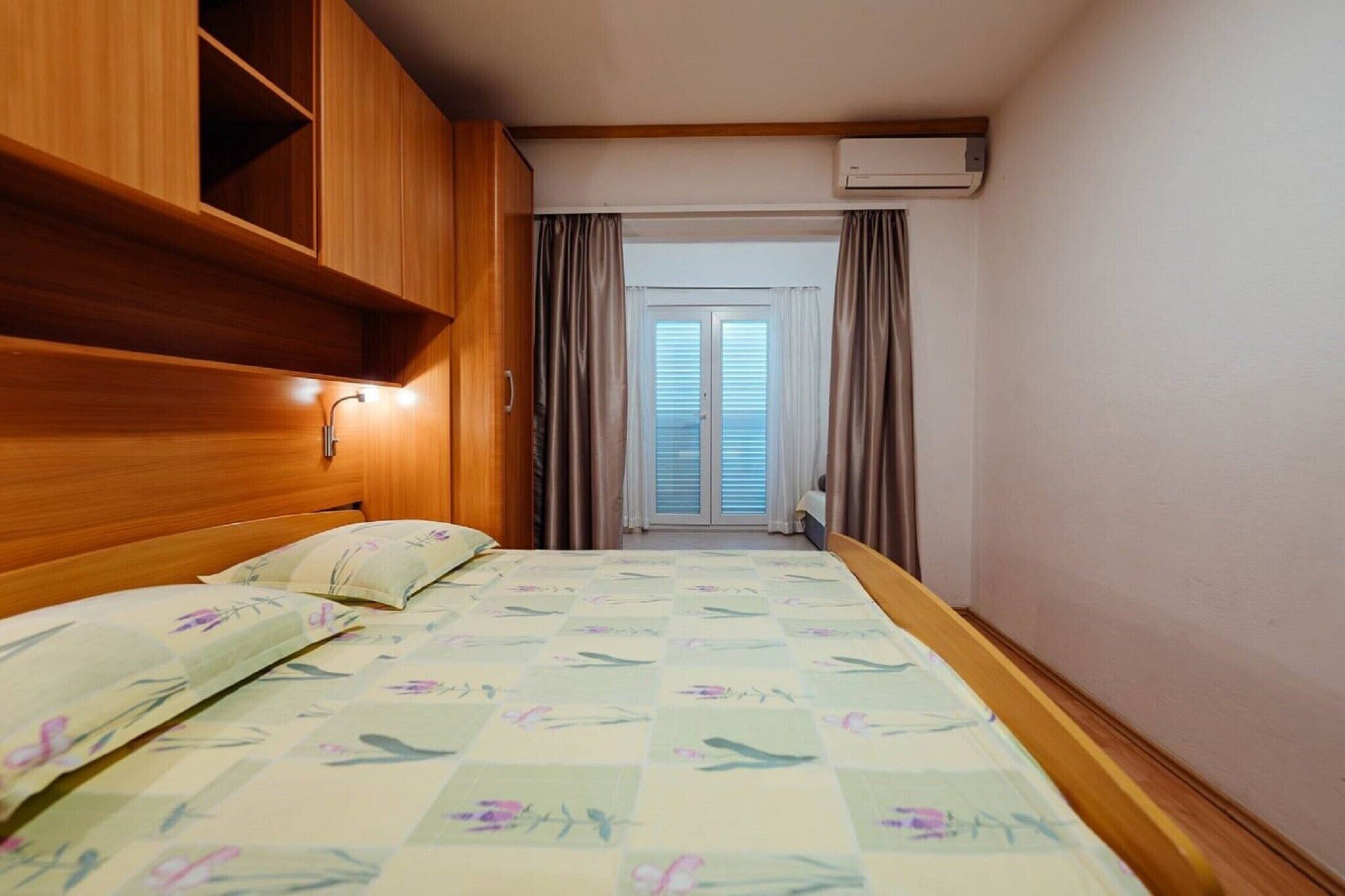 A bedroom with a wooden headboard, a bed with floral bedding, a window with curtains, and an air conditioning unit.