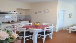 A dining area with a round table, white chairs, a clock on the wall, and a kitchen in the background. - Apartments Jezerane - Studio Apartment (Jezerane)