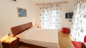Une chambre à coucher avec un lit, une table de chevet avec une lampe, un pouf rouge, un téléviseur fixé au mur et une fenêtre avec des rideaux à motifs.