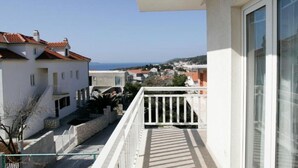 Un balcon donnant sur une ville côtière, un bâtiment blanc avec un toit recouvert de tuiles rouges et un ciel dégagé.