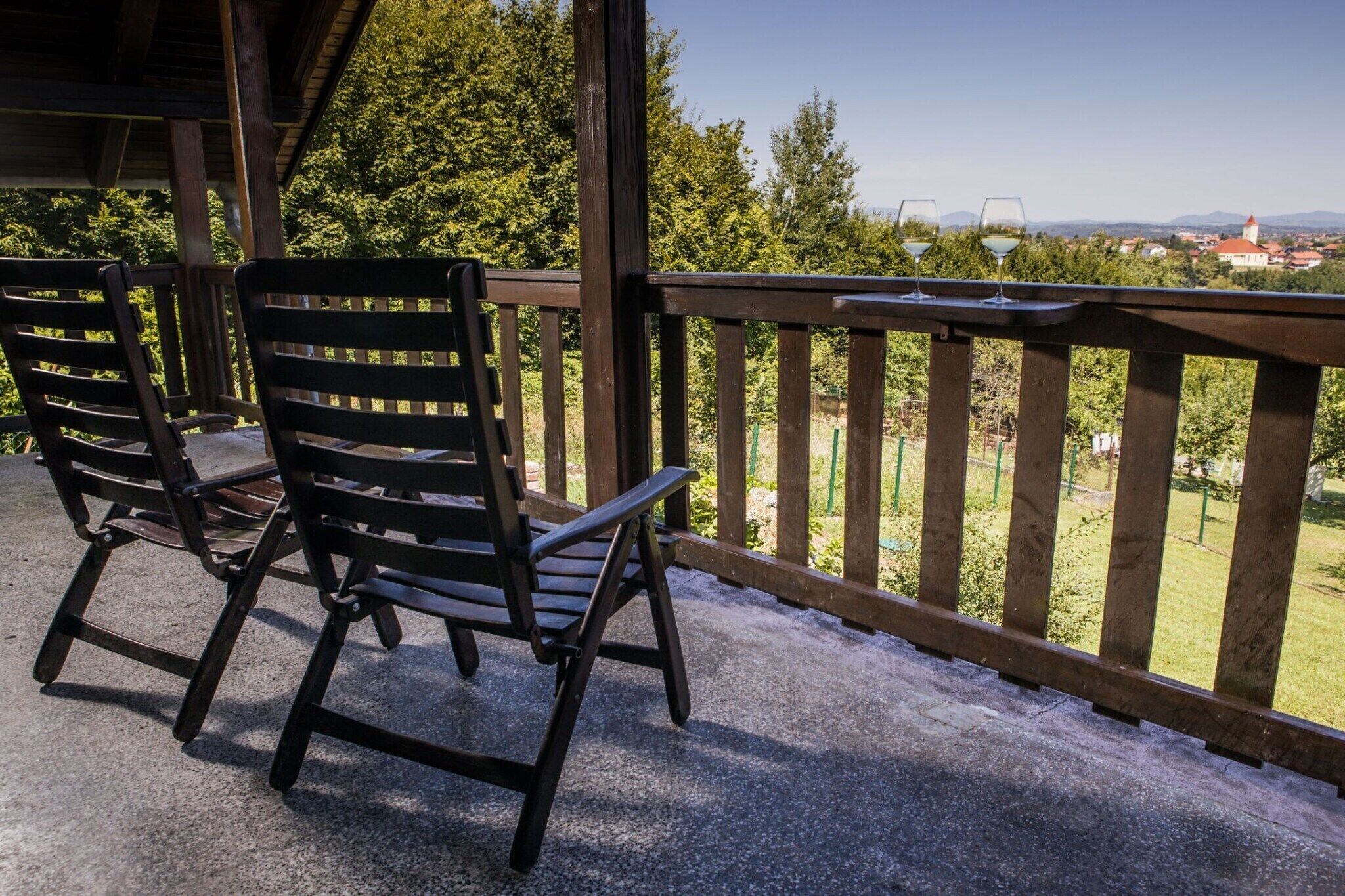 A wooden deck with two chairs and a table, overlooking a scenic landscape with trees and distant buildings.