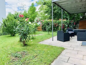 A covered patio with outdoor furniture, a garden with various plants and flowers, and a wooden fence in the background.