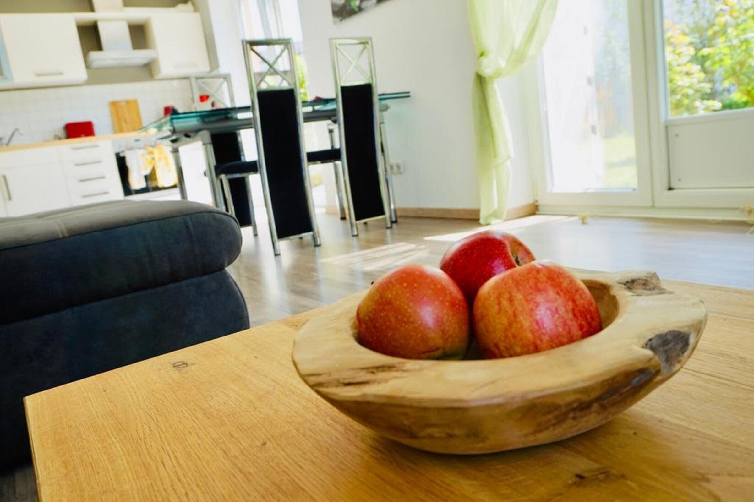 En träskål med äpplen står på ett bord i ett modernt kök med vita skåp och en matplats.