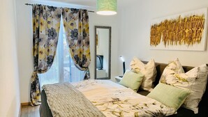 Ein Schlafzimmer mit einem Bett, Kissen, einem Nachttisch mit Lampe, einem Spiegel und einem Fenster mit Vorhängen.