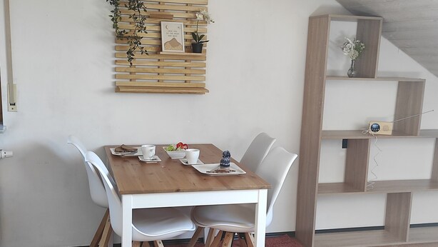 A dining area with a wooden table, white chairs, a small shelf with plants, and a wall-mounted shelf with a picture.