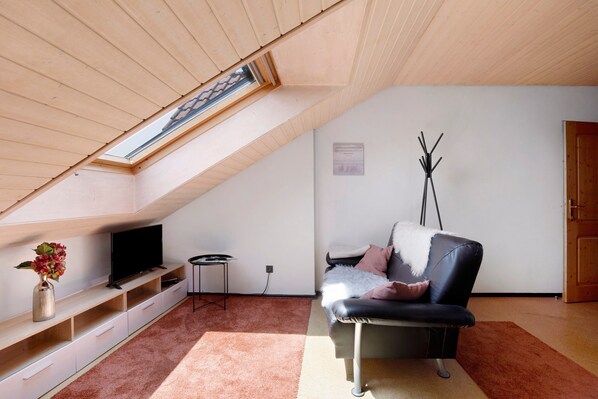 Ein modernes Wohnzimmer mit schrägverdeckter Decke, einem Flachbildfernseher, einem Sofa, einem kleinen Beistelltisch und einer Vase mit Blumen.