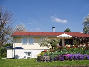 Uma casa de dois andares com telhado vermelho, varanda coberta e jardim com diversas flores.