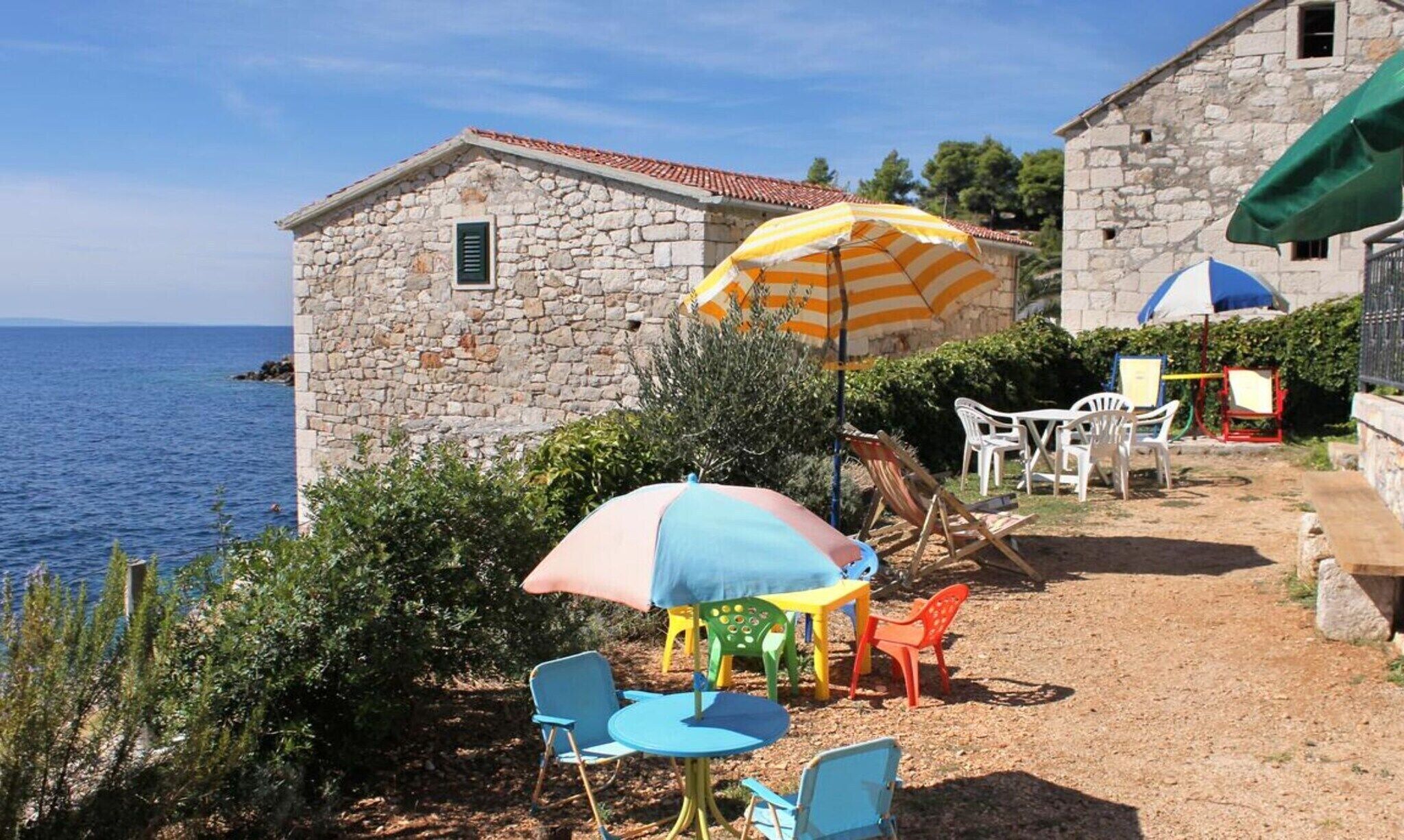 Un bâtiment en pierre avec un toit en tuiles, des places assises à l’extérieur avec des chaises colorées et un parasol à rayures, et une vue sur la mer.