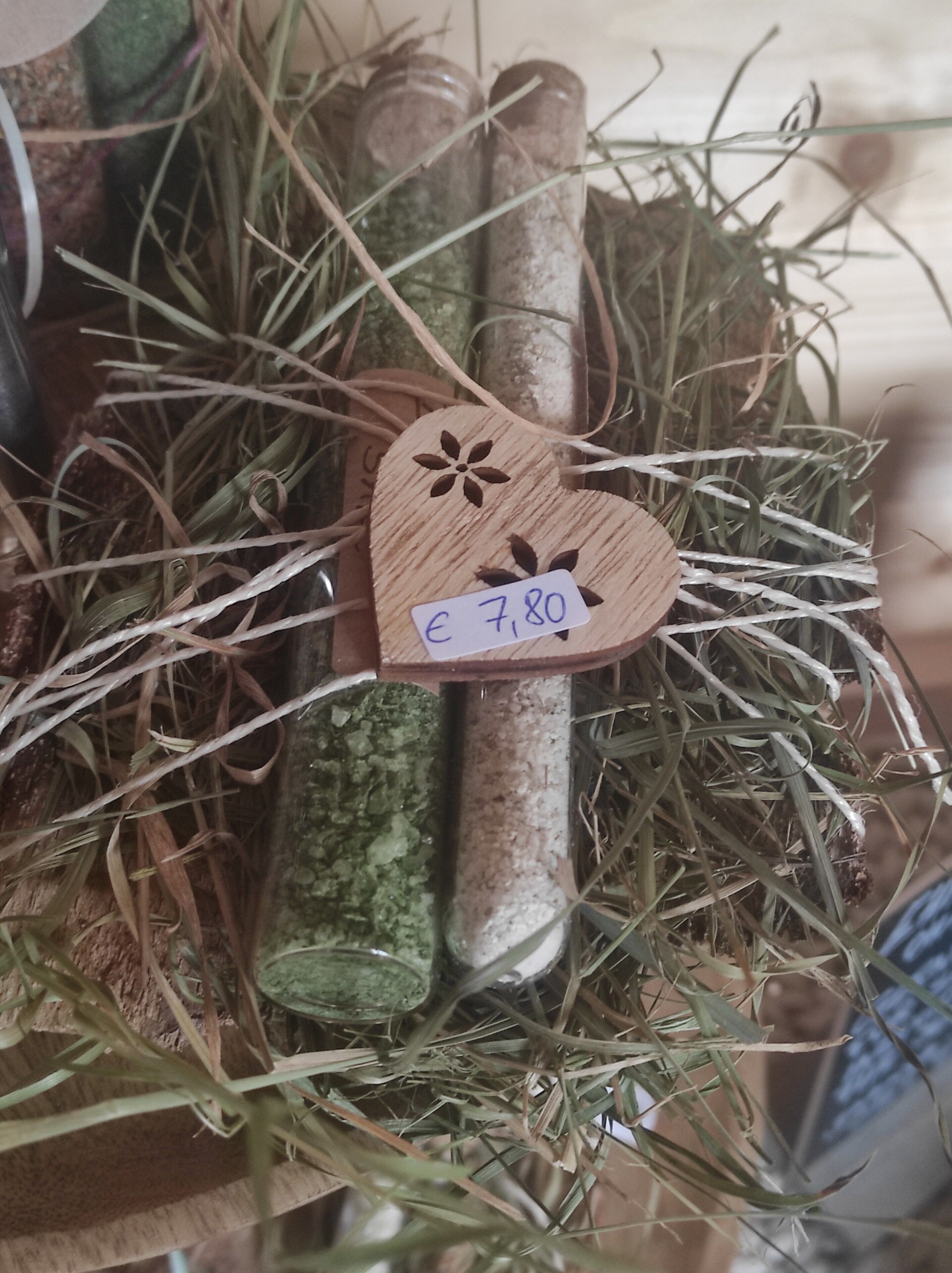 Un petit objet décoratif en bois en forme de cœur, muni d’une étiquette indiquant son prix, entouré d’herbe séchée et de feuilles vertes.