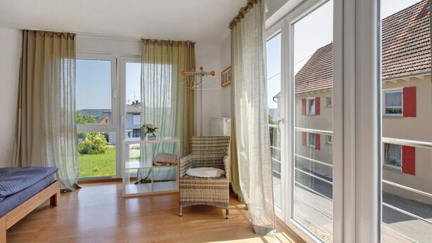 Una habitación con una cama, una silla de mimbre, una mesa con un florero y vista a un edificio a través de una ventana.