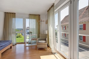 A room with a bed, a wicker chair, a table with a vase, and a view of a building through a window.