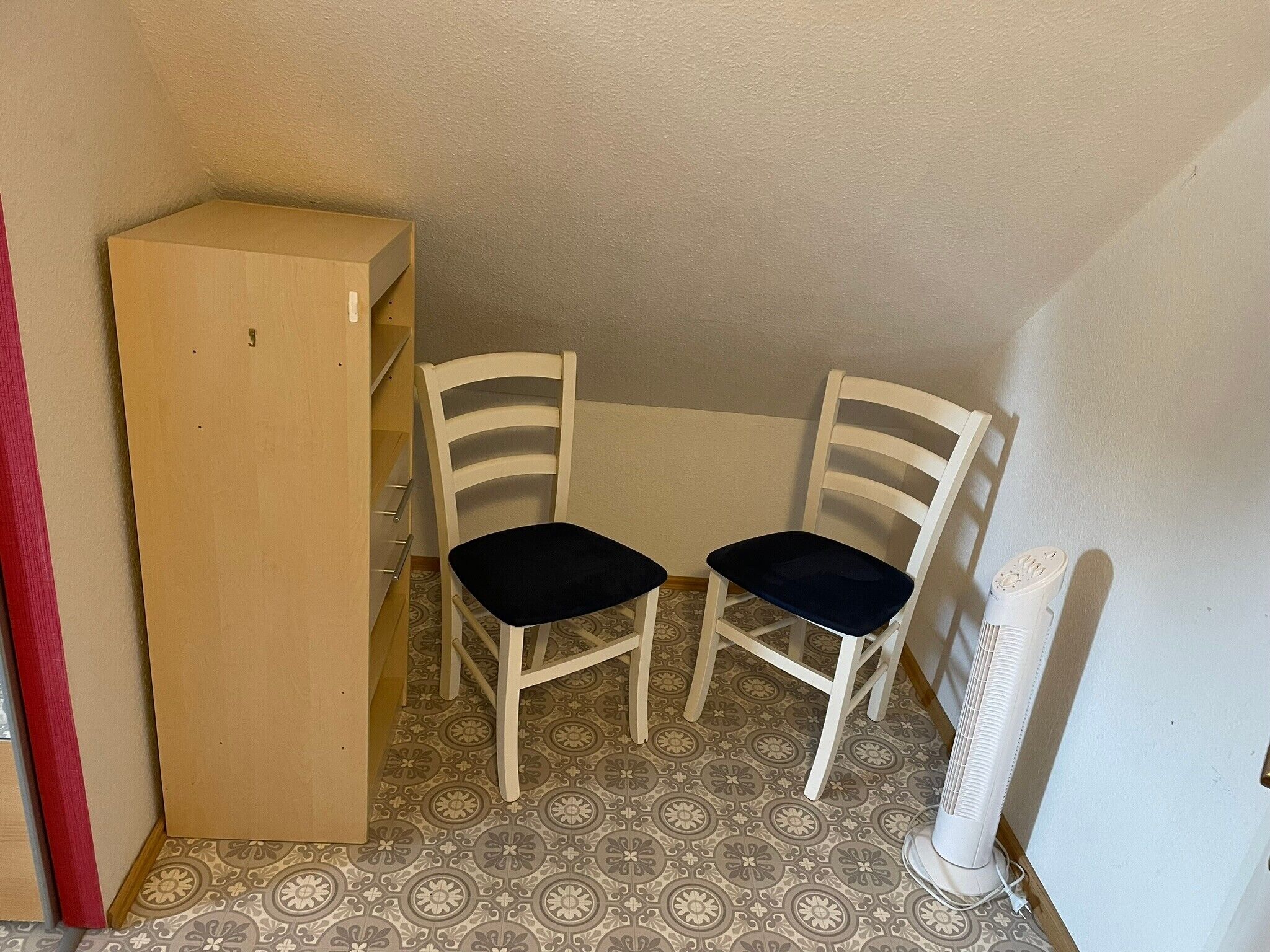 A corner room with two white chairs, a wooden cabinet, and a standing fan.