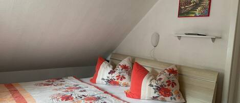 A bedroom with a sloped ceiling, a bed with floral bedding, a painting of a garden, and a bedside table.