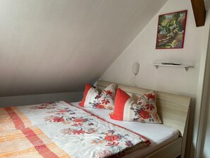 Une chambre avec un plafond en pente, un lit recouvert d’une literie à motifs floraux, un tableau représentant un jardin et une table de chevet.