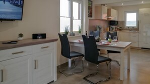 A modern kitchen with white cabinets, a dining table with chairs, a TV mounted on the wall, and a window providing natural light.