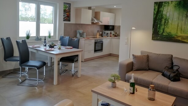 A modern kitchen with white cabinets, a dining area with a table and chairs, and a living room with a sofa and a painting.