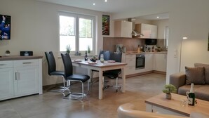 Eine moderne Küche mit weißen Schränken, eine Essecke mit Tisch und Stühlen, ein Sofa und ein an der Wand befestigter Fernseher.