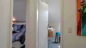 Ein Flur, der zu einem Schlafzimmer mit Bett sowie zu einem Badezimmer mit Waschbecken und Stuhl führt.