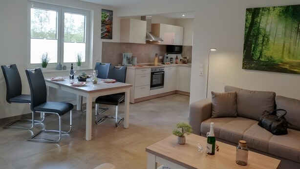 Eine moderne Küche mit weißen Schränken, ein Essbereich mit Tisch und Stühlen, eine Couch und ein Gemälde von einem Wald.