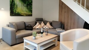 Ein modernes Wohnzimmer mit einem grauen Sofa, einem weißen Couchtisch und einem weißen Sessel. Eine Treppe mit Holzwandungen ist rechts zu sehen.