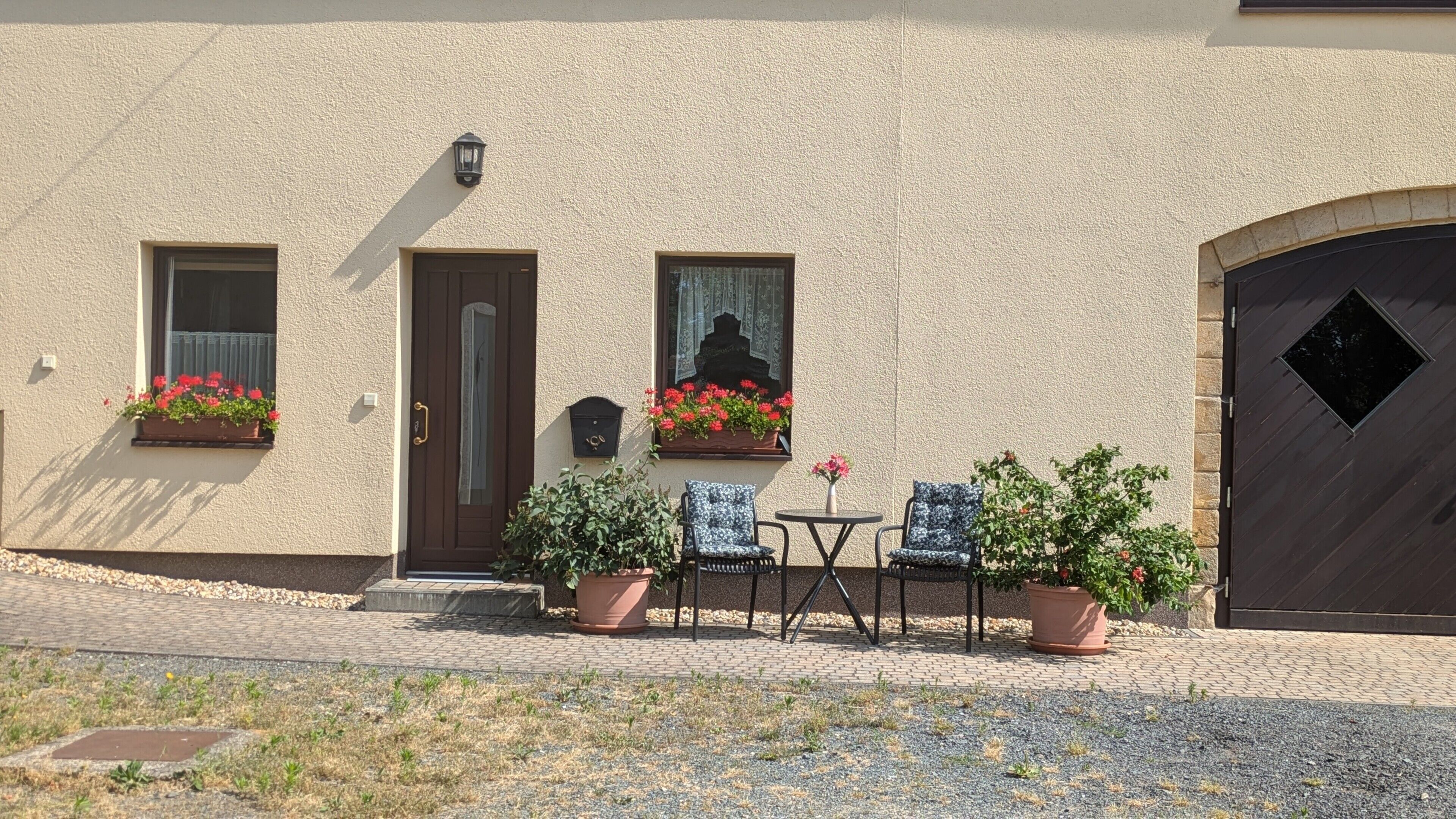 Ein Gebäude mit beige-farbener Fassade, zwei Fenstern mit Blumenkästen, einer Tür und einem kleinen Tisch mit Stühlen.