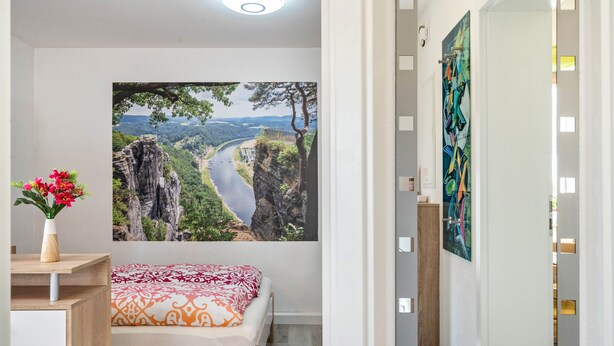 Un dormitorio con cama, escritorio, un florero con flores y un cuadro de paisaje grande en la pared.