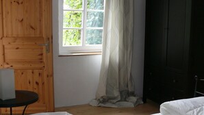 Um quarto com uma cama, porta de madeira, janela com cortinas e uma pequena mesa.