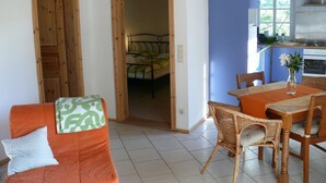 Une pièce avec une porte en bois, un lit recouvert d’une couverture à motifs verts et blancs, une chaise en osier, une table en bois avec un vase, et un coin cuisine équipé d’une cuisinière et d’un évier.
