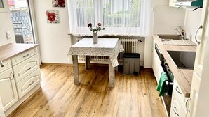 Una cocina compacta con suelo de madera, una mesa con mantel de encaje, un radiador y una ventana con cortinas de gasa.