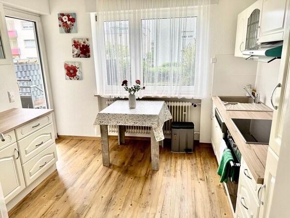 A compact kitchen with a wooden floor, a table with a lace tablecloth, a radiator, and a window with sheer curtains.