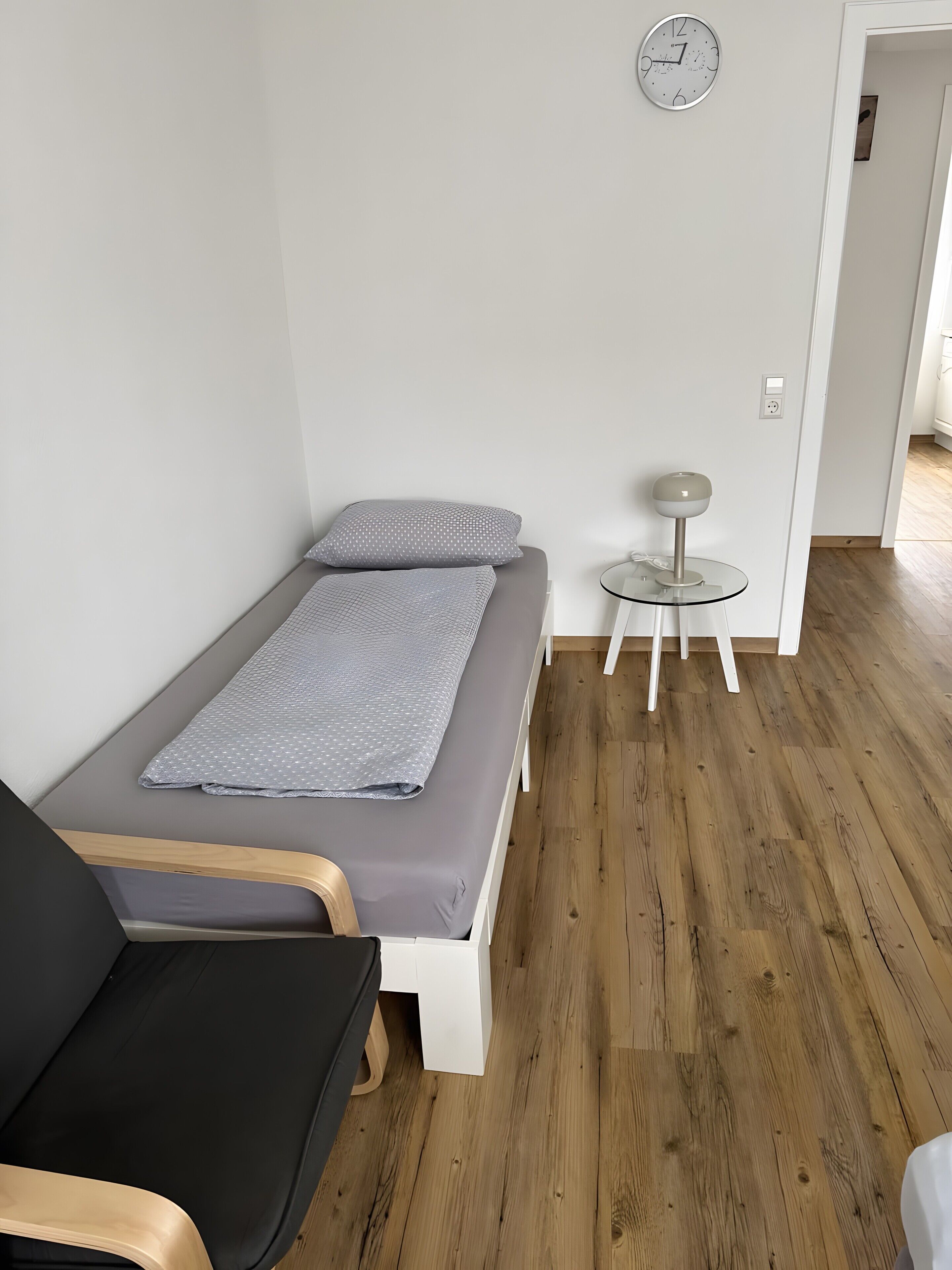 Une petite chambre avec un lit simple, un plancher en bois, une table de chevet avec une lampe et une horloge au mur.