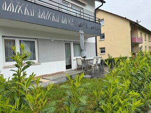 Een modern gebouw met balkon, een eetgedeelte met stoelen en tafel, en een tuin met groene planten.