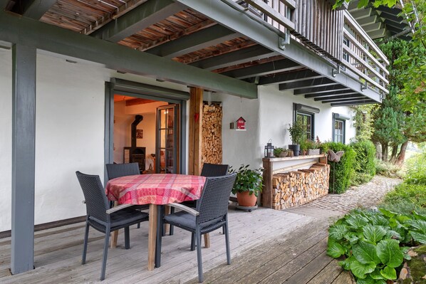 Ein überdachter Außenbereich mit Tisch, Stühlen, einem Holzstapel und einem Gartenweg.