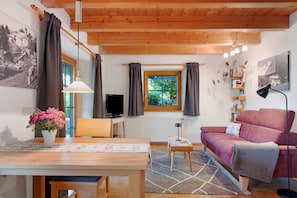 Un salon chaleureux avec un plafond en bois, une table à manger agrémentée d’un vase de fleurs, un canapé, un téléviseur et une fenêtre avec des rideaux.