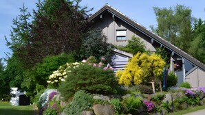 Una casa con techo inclinado, rodeada de un jardín con diversas plantas y flores.