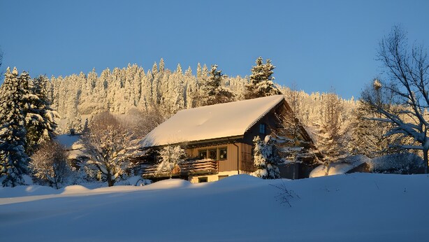 Una cabaña de troncos con techo cubierto de nieve, rodeada de árboles cargados de nieve y un cielo azul despejado.