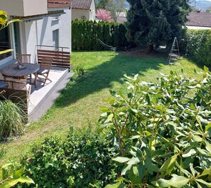 Un patio con mesa y sillas, un pequeño jardín con diversas plantas y un seto al fondo.